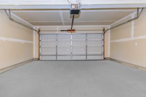 An empty garage interior with a concrete floor and a closed silver garage door. The walls are painted beige, and there are unfinished patches on the drywall. A ceiling-mounted garage door opener is visible above the door. The space is well-lit and uncluttered.