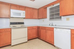 A kitchen featuring light wooden cabinets, a white microwave above a stove, and a dishwasher. The countertops are light-colored, and there's a sink beneath a window. The flooring is a beige tile, creating a warm, inviting atmosphere. Natural light is coming through the window.