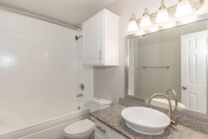 A modern bathroom featuring a white bathtub, a granite countertop with a round sink, a toilet, and a mirrored cabinet with overhead lighting. The walls are painted in a light color, and there is a towel bar next to the doorway. The overall design is clean and contemporary.