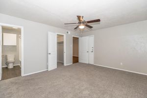 A spacious, well-lit room featuring light gray walls and a ceiling fan. Two white panel doors lead to separate compartments, one likely a closet and the other a bathroom. The floor is covered in soft gray carpet, creating a cozy atmosphere.