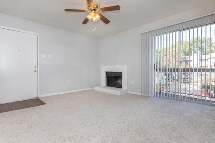 An empty living room with light-colored carpet, a ceiling fan with wooden blades, and a white fireplace. A sliding glass door with vertical blinds provides natural light and a view of the outdoors. The walls are painted a light gray, creating a spacious and airy atmosphere.