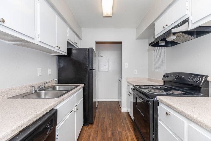 A modern kitchen featuring white cabinets, a black refrigerator, an electric stove, and a spacious countertop. The walls are painted light gray, and the floor has a wood laminate finish. A door is visible at the end of the kitchen, leading to another area. Bright overhead lighting illuminates the space.
