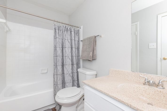 A clean and bright bathroom featuring a white bathtub with a shower curtain, a toilet, a beige countertop with a sink, and a mirror above the sink. The walls are painted in a light gray color, and there is a towel hanging on a rack. The floor is wooden.
