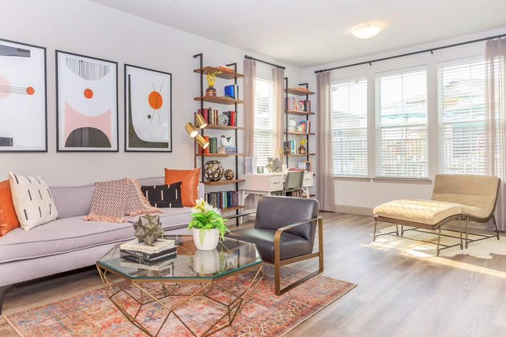 A modern living room featuring a light gray sofa with orange and white cushions, a glass coffee table, and a decorative rug. There are framed art pieces on the wall, a bookshelf filled with books, and a desk by the window. Natural light filters through sheer curtains, creating a bright and inviting atmosphere.