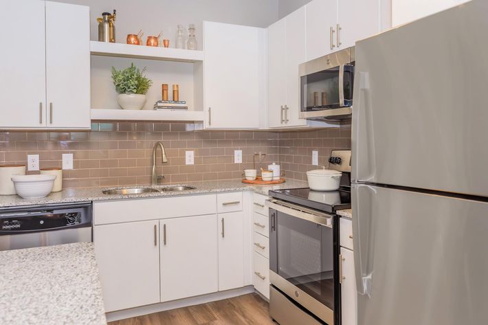 Modern kitchen featuring white cabinets, a stainless steel fridge, and an oven. The countertop is made of speckled granite, with a bowl and decorative items on display. The backsplash is a warm, neutral tile, and there are plants and glassware on the shelves above the sink. The space is bright and inviting.