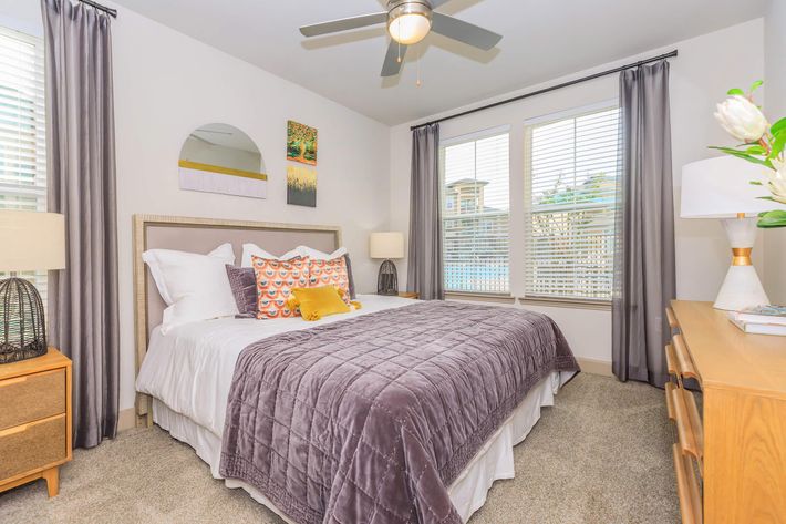 A cozy bedroom with a king-sized bed dressed in white and gray linens, accented with colorful pillows. There's a bedside table with a lamp, a modern dresser, and large windows with sheer curtains allowing natural light. A ceiling fan and a decorative wall mirror add to the inviting atmosphere.