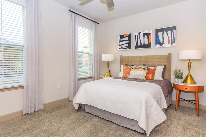 A cozy bedroom featuring a neatly made bed with decorative pillows, two stylish bedside lamps, and artwork on the wall. Large windows with light curtains allow natural light to brighten the space. The carpeted floor adds warmth, and a small table beside the bed holds a plant for a touch of greenery.