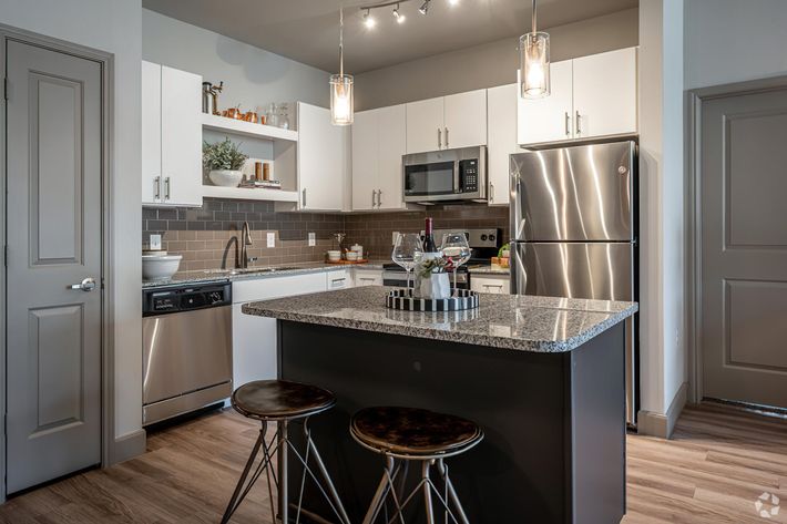 A modern kitchen featuring white cabinetry, stainless steel appliances, and gray tile backsplash. The island has a granite countertop with two bar stools, and a small decorative centerpiece. Stylish pendant lights hang above the island, creating a warm and inviting atmosphere.