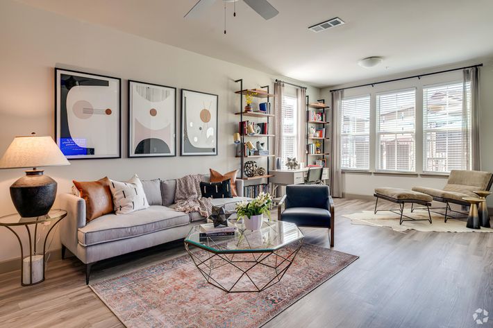 A modern living room featuring a light gray sofa adorned with colorful cushions, a glass coffee table, and stylish artwork on the walls. A sleek lamp stands beside the sofa. There's a bookshelf filled with books and decorative items, alongside large windows that provide natural light, and a cozy armchair in the corner.