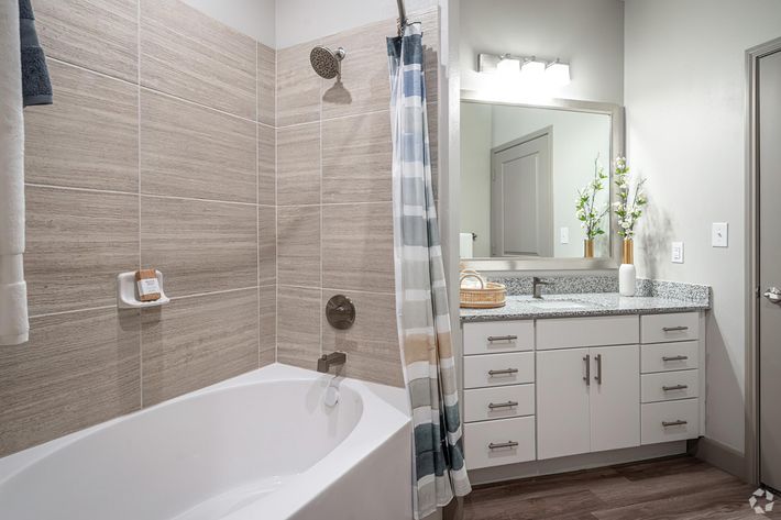 A modern bathroom featuring a bathtub with a shower, light-colored tiled walls, and a mirror with bright lighting. The vanity has white drawers and a countertop with a decorative basket. A shower curtain with a striped pattern hangs nearby, and there are some green plants on the countertop for a fresh look.