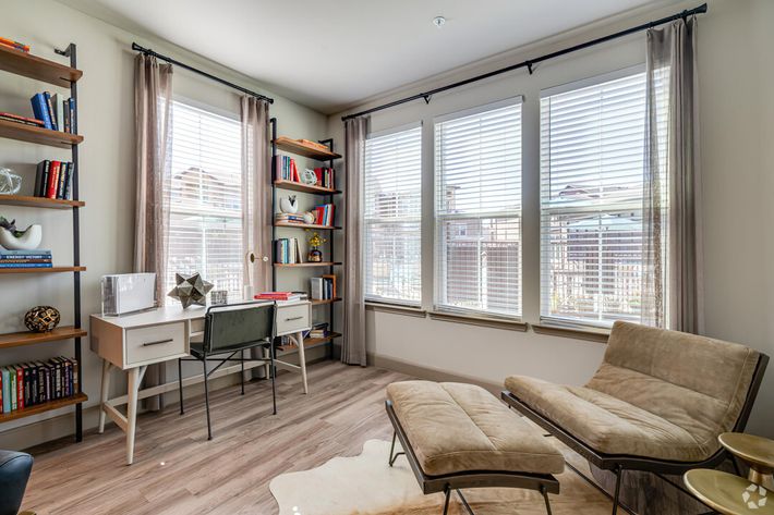 A bright, stylish home office featuring large windows with sheer curtains, a light wooden desk with a chair, a bookshelf filled with books and decorative items, and a cozy lounge chair on a plush area rug. Natural light fills the space, creating a warm and inviting atmosphere.