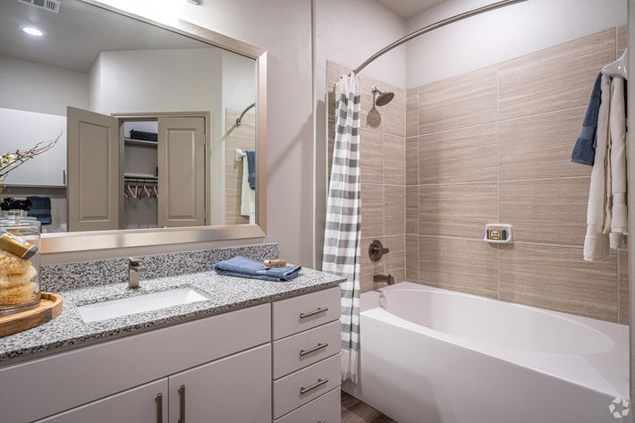A modern bathroom featuring a white bathtub, a shower with a glass curtain, a granite countertop sink with a mirror above. There are light-colored tiles on the walls, and storage cabinets with folded towels. A decorative vase and blue towels add a cozy touch to the space.