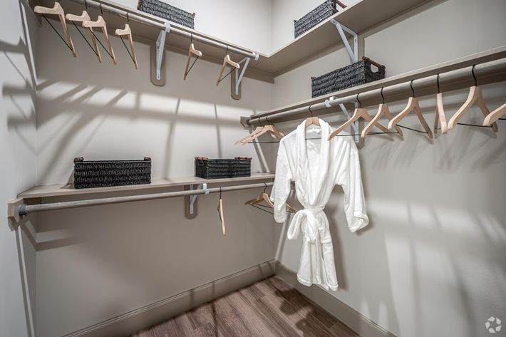 A well-organized closet featuring wooden hangers, a white bathrobe hanging on a rail, and neatly arranged baskets on shelves. The walls are painted a light color, and the floor is made of wood, creating a modern and spacious feel. Natural light casts soft shadows across the room.