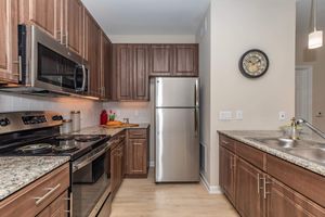 A modern kitchen featuring wooden cabinetry, stainless steel appliances including a refrigerator and oven, granite countertops, and a sink. The space is well-lit with natural light and has a wall clock for added decor. Red kitchen accessories add a splash of color to the neutral tones.