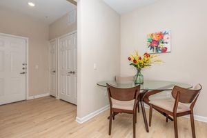 A modern dining area featuring a round glass table with three wooden chairs. A vase filled with colorful flowers sits in the center of the table. The wall beside the table displays a small floral painting. Nearby are closed white sliding doors and an entryway with a light beige wall.