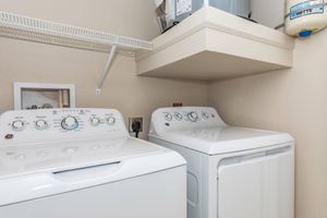 A laundry room featuring a white washing machine and a white dryer side by side. Above, there is a shelf with storage items. The wall is light-colored, and there is a small framed picture on the left wall. The appliances have control dials and buttons visible on their fronts.