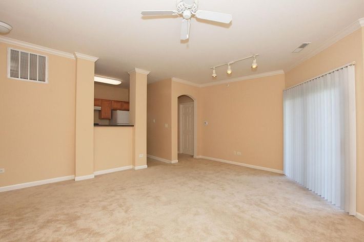 An empty living room featuring beige walls and carpeting, with a ceiling fan and track lighting. An archway leads to a kitchen area with wooden cabinets visible in the background. A sliding glass door opens to an exterior space, and a small wall vent is located to the left.