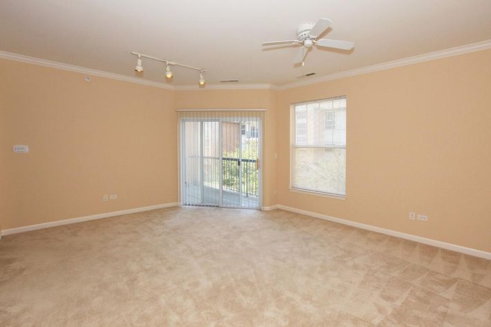 A spacious, empty living room with light beige walls and tan carpet. The room features a ceiling fan and recessed lighting. There's a large sliding glass door leading to a balcony, with a view of greenery outside. Natural light streams in through the window beside the door, enhancing the open feel of the space.