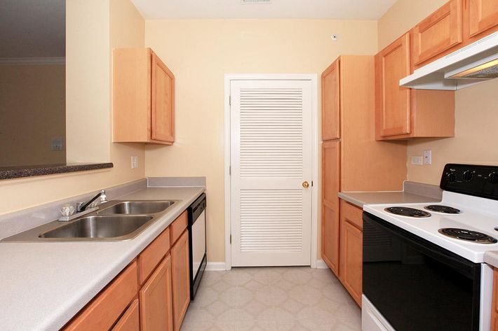 A kitchen featuring light wood cabinets, a double sink, a black dishwasher, and a white stove with a built-in oven. The walls are painted a soft yellow, and there is a door leading to another room. The countertop is a neutral color, and the flooring has a subtle pattern.