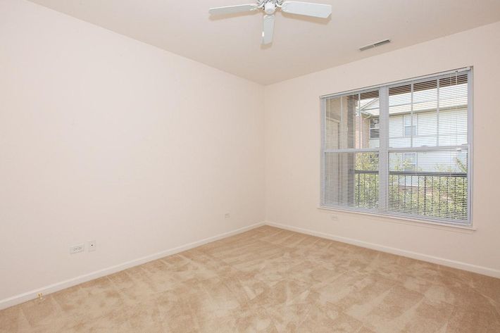 Empty room featuring a light-colored carpet and beige walls. A window with blinds allows natural light to enter, revealing a view outside. A ceiling fan is mounted above. The overall ambiance is bright and spacious, ideal for potential decorating or use as a living space.