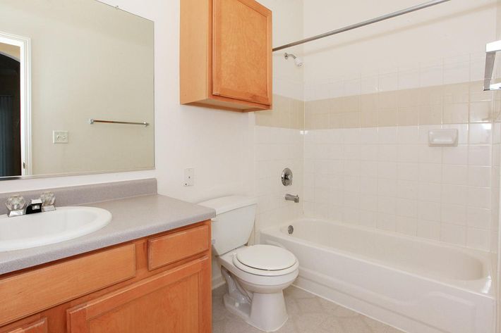 A clean and simple bathroom featuring a bathtub with a shower, a white toilet, a sink with a countertop, wooden cabinetry, and a mirror. The walls are tiled with light-colored ceramic tiles, and the flooring is beige. Natural light is coming from an unseen window.
