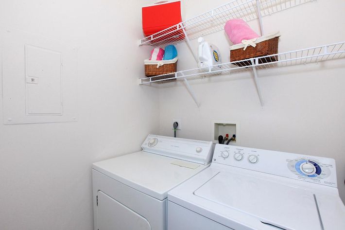 A laundry room featuring a white washing machine and dryer side by side. Above, a wire shelf holds colorful baskets, including pink items and laundry detergent. The walls are light-colored, and there is a small panel on the left side.