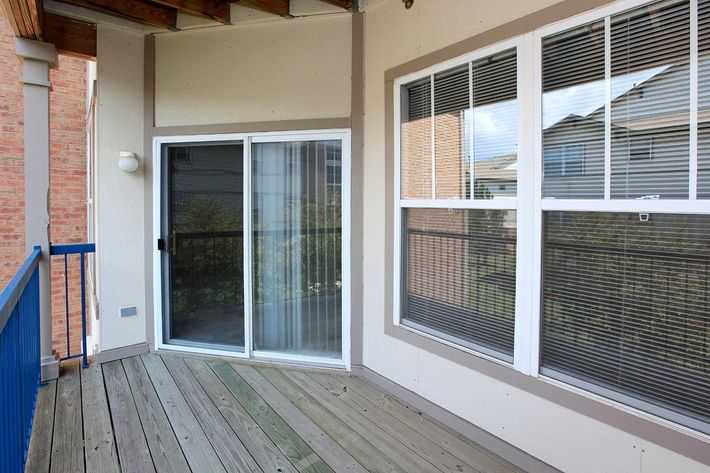 A view of a balcony featuring a sliding glass door, wooden flooring, and large windows with square grids. The balcony is bordered by a blue railing and overlooks a residential area with buildings visible in the background. Natural light fills the space, creating a bright and open atmosphere.