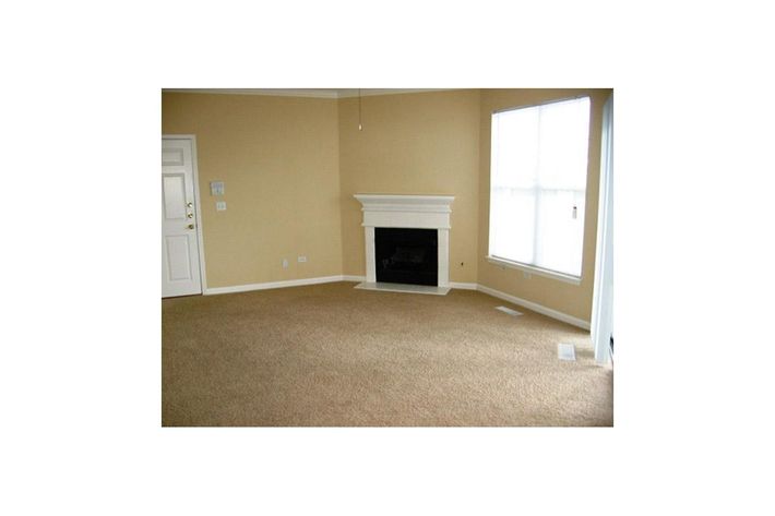 A spacious, empty living room featuring beige walls and carpet. It includes a white fireplace against one wall, a large window allowing natural light, and a door on the left. The room is unfurnished, emphasizing its openness and potential for decoration.