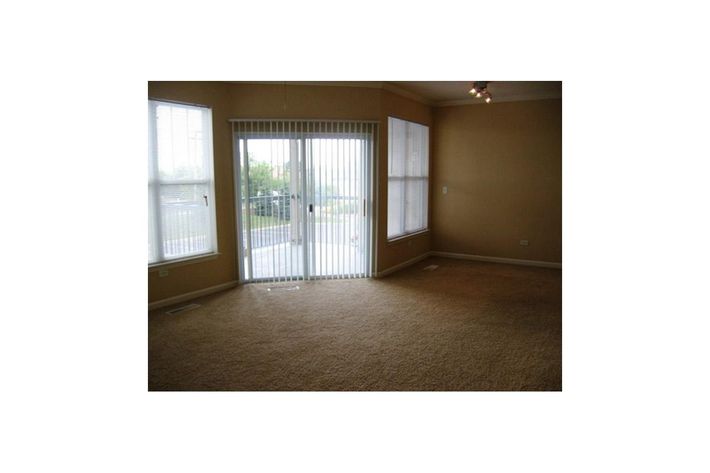 A spacious empty room with light beige walls and carpet, featuring large windows with vertical blinds. One wall has a sliding glass door that leads outside, while the other two walls have smaller windows allowing natural light. The ceiling has a simple light fixture.