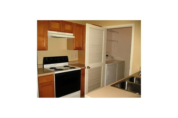 A small kitchen featuring wooden cabinetry, a white stove with four burners, and a double sink. An open door leads to a laundry area with shelves and a washer/dryer unit. The walls are painted a light color, creating a warm and inviting atmosphere.