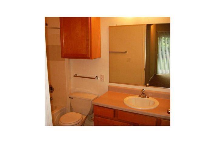 A compact bathroom featuring a white toilet, a sink with a faucet, and a wooden vanity. There is a mirror above the sink and a shower area visible. Light wood cabinets are mounted above the sink, and the room is well-lit with a window in the background showing a view of an outside area.