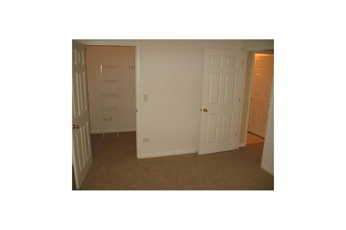A simple room with beige carpet and light-colored walls. Two sets of double doors are visible, leading to different areas, one with a small shelving unit visible. The room is well-lit and empty, giving a sense of space and potential for use as a bedroom or a small office.