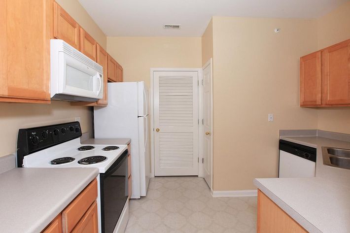 A modern kitchen featuring light wood cabinets, a white refrigerator, a black stove, a microwave above the stove, and a dishwasher. The countertops are gray, and the room has beige walls with a door leading to a closet or pantry. The layout is open and functional, designed for everyday use.