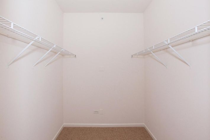 Empty closet with white wire shelves on both sides. The walls are painted white, and there is beige carpet on the floor. The space appears clean and uncluttered, suitable for storage or organizing items.