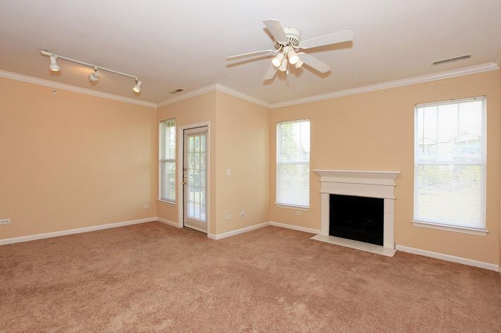 Empty living room with beige walls and brown carpeting. Features include a ceiling fan, track lighting, two windows with blinds, and a fireplace with a white mantel. A door leads outside, providing natural light to the space.