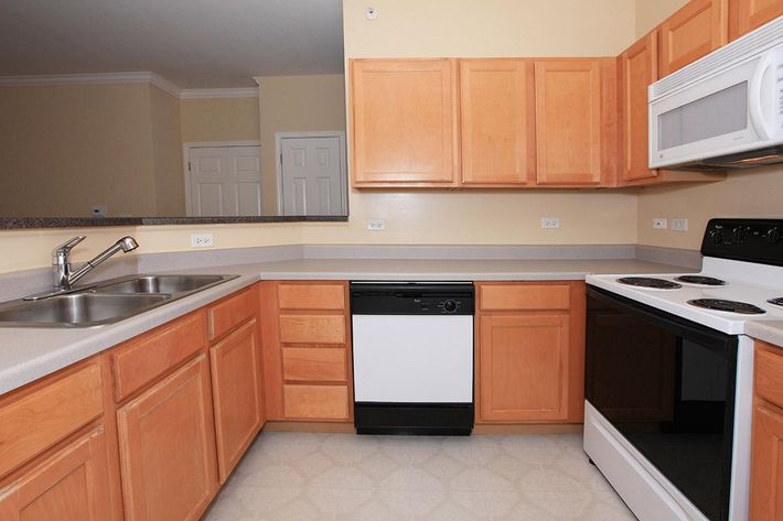 A modern kitchen featuring light wood cabinets, a double sink, a black dishwasher, a white microwave, and a black stove. The countertops are neutral in color, and there's a mirrored backsplash. The floor is light-colored vinyl tiles, and the space has a minimalistic design with adequate kitchen appliances.