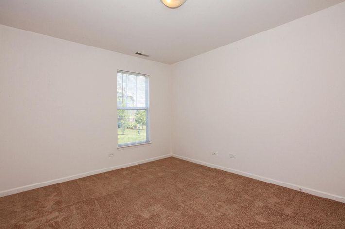 A vacant room featuring light-colored walls, a window letting in natural light, and a carpeted floor. The space appears empty with no furniture or decorations, emphasizing its openness and potential for customization.