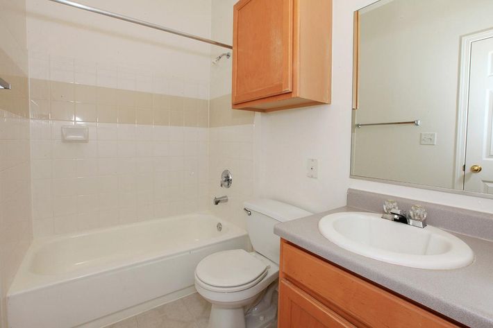 A small bathroom featuring a bathtub with a shower, a toilet, and a sink with a countertop. Wooden cabinets above the sink provide storage. A large mirror hangs above the sink, and the walls are light-colored tiles with a simple layout. The flooring is a light tile pattern.
