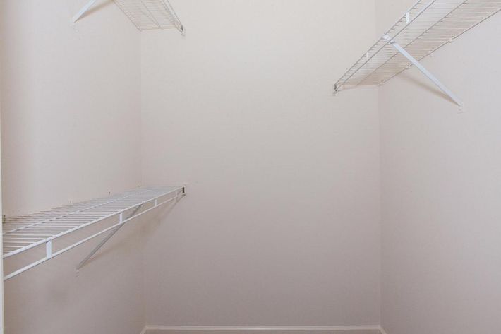 A spacious, empty closet with white wire shelving installed on the left wall and back wall. The walls are painted a light color, creating a clean, minimalistic look. The floor is bare, emphasizing the uncluttered state of the space.