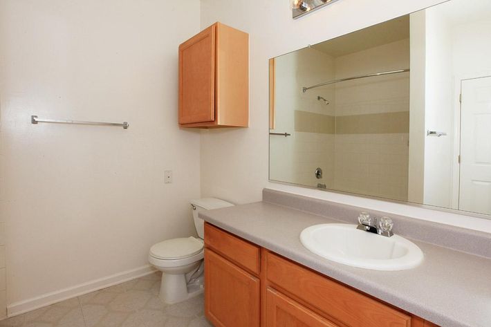A clean, spacious bathroom featuring a sink with two faucets, a large mirror above it, wooden cabinetry, a toilet, and a shower area with glass doors. The walls are painted in a light color, and the flooring is laid with light tiles. A towel rack is mounted on one wall.