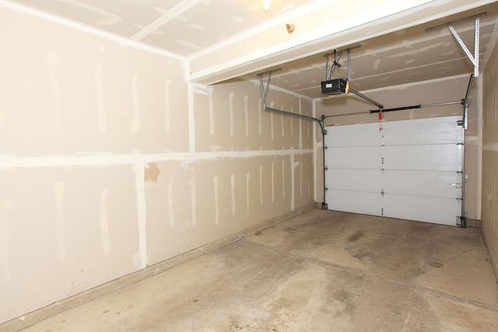 A vacant garage with unfinished drywall, concrete floor, and a closed white garage door. The walls have visible markings and patches, and there are no vehicles or furniture present in the space. Overhead, there is a garage door opener mounted on the ceiling.