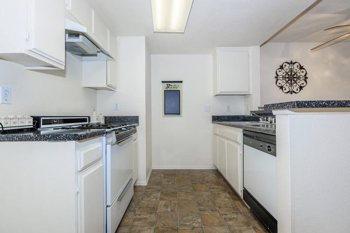 A modern kitchen featuring white cabinetry, stainless steel appliances including an oven and dishwasher, and a dark granite countertop. The floor has a tiled design, and there is a wall-mounted decorative piece. Natural light illuminates the space, providing a clean and inviting atmosphere.