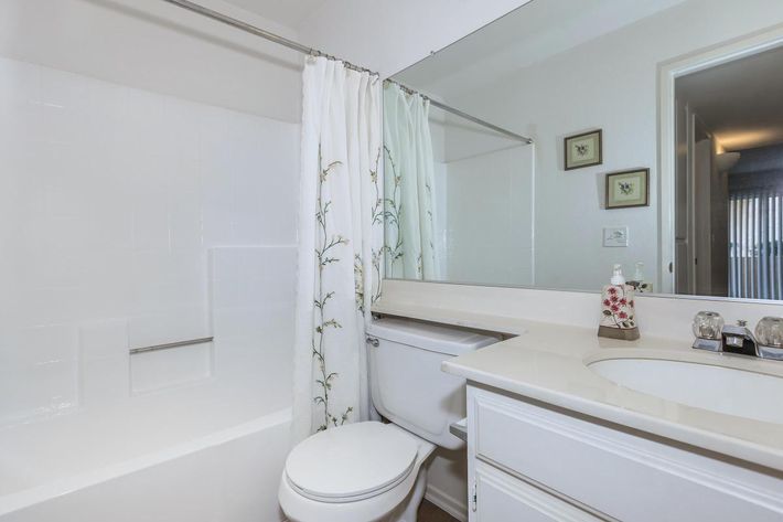 A clean and modern bathroom featuring a white bathtub with a shower curtain adorned with floral designs, a toilet, and a light-colored vanity with a sink. The walls are painted in soft tones, and two framed pictures are hung above the vanity. Natural light enters through an adjacent room.