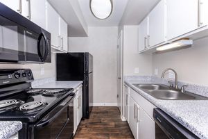 A modern kitchen featuring a black stove, microwave, and refrigerator, with white cabinets and a granite countertop. The space has a sink with a chrome faucet and wood-like flooring, illuminated by overhead lighting, creating a bright and functional cooking area.
