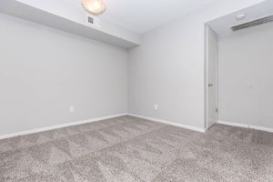 Empty room featuring light gray walls and a carpeted floor. A ceiling light is visible, and there is a door on the right leading to another space. The lighting creates minimal shadows, highlighting the spaciousness of the area. No furniture or decorations are present.