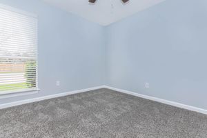 An empty room with light blue walls and a large window featuring white blinds. The floor is covered with grey carpet. There is a ceiling fan with wooden blades, and the room appears well-lit with natural light coming through the window.