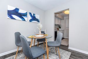 A modern dining area featuring a glass table set for four with gray chairs. Decorative accents include a placemat and wine glasses. Two abstract blue paintings adorn the wall above the table, and a glimpse of a kitchen is visible through an open doorway. The floor is covered with a light-colored rug.