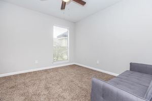 A minimalist room featuring beige carpet and light gray walls. A small window with blinds lets in natural light. A gray sofa is positioned against one wall, creating a simple and uncluttered space.