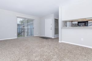 A modern, unfurnished living space featuring light gray walls, a beige carpet, and large windows with vertical blinds. A door leads outside on the left, while a kitchen area with a countertop and a microwave is visible in the background. The overall ambiance is bright and airy.