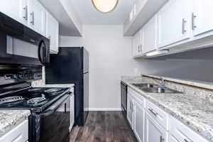 Modern kitchen featuring white cabinetry, black appliances including a stove, microwave, and refrigerator, and a granite countertop with a double sink. The flooring is a dark wood finish, and the space is well-lit with a ceiling light.